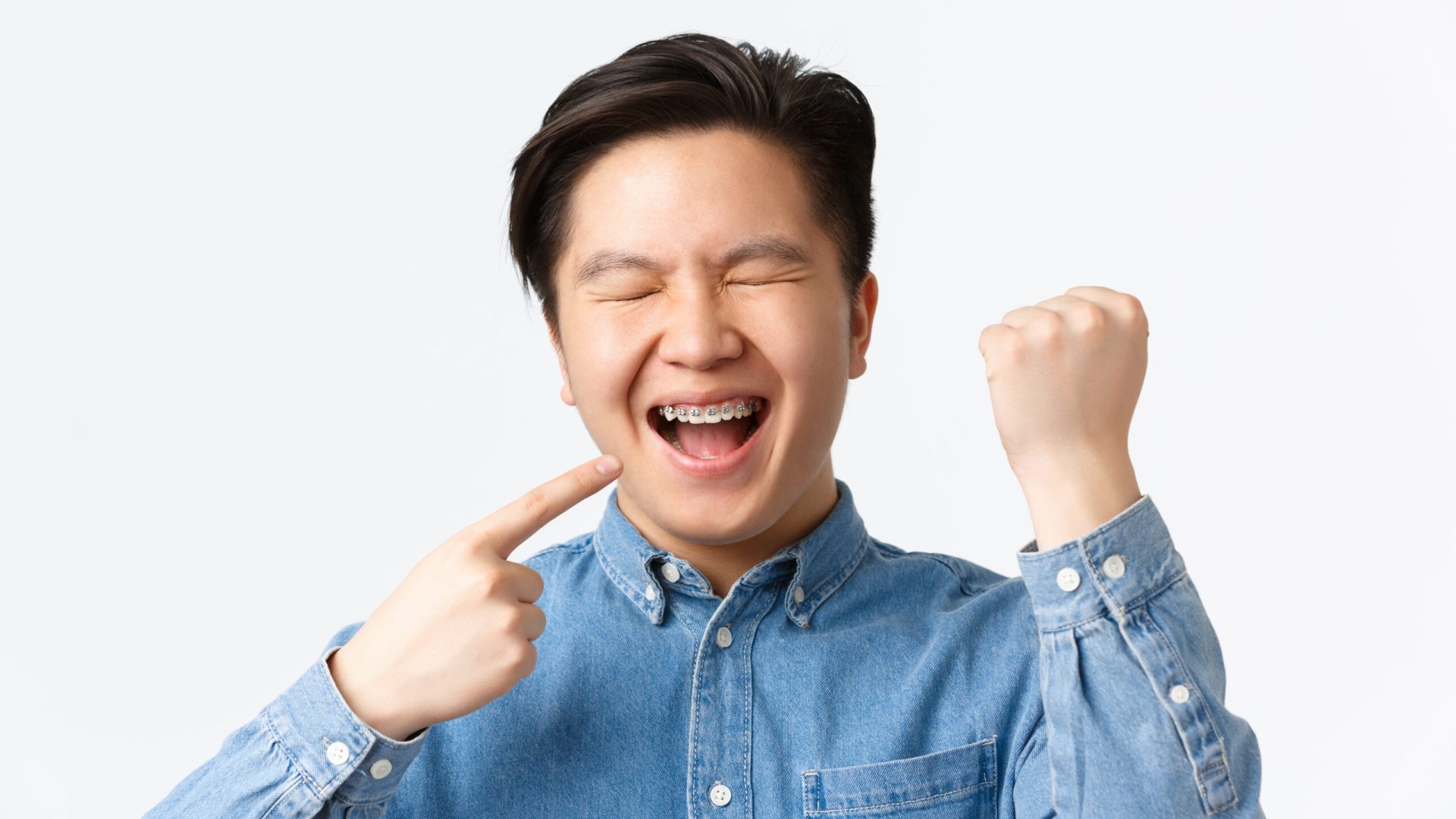 Orthodontics and stomatology concept. Close-up of excited and happy asian guy rejoicing over new teeth braces, pointing at mouth and smiling, fist pump, triumphing over white background.